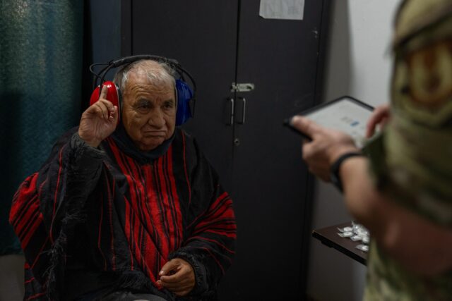 An older adult wearing traditional red-and-black clothing participates in a hearing test while seated. Wearing a red-and-blue WAHTS headset, the individual raises one finger to indicate hearing a tone while a clinician records responses on a tablet. The image demonstrates portable hearing testing in a community or field healthcare setting.