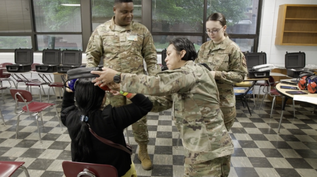 A military service member fits a WAHTS headset on a seated participant while two other uniformed service members observe during a group hearing testing session.